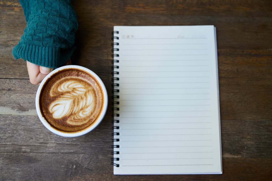 Flat lay of a coffee cup with latte art next to a blank notebook on a wooden table.