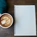 Flat lay of a coffee cup with latte art next to a blank notebook on a wooden table.