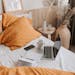 From above of open netbook with black screen and cup of coffee with smartphone and notebook placed on crumpled bed sheet in bedroom with big vase on floor