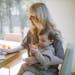 A mother works on a laptop at home while holding her baby, showcasing remote working and parenting.