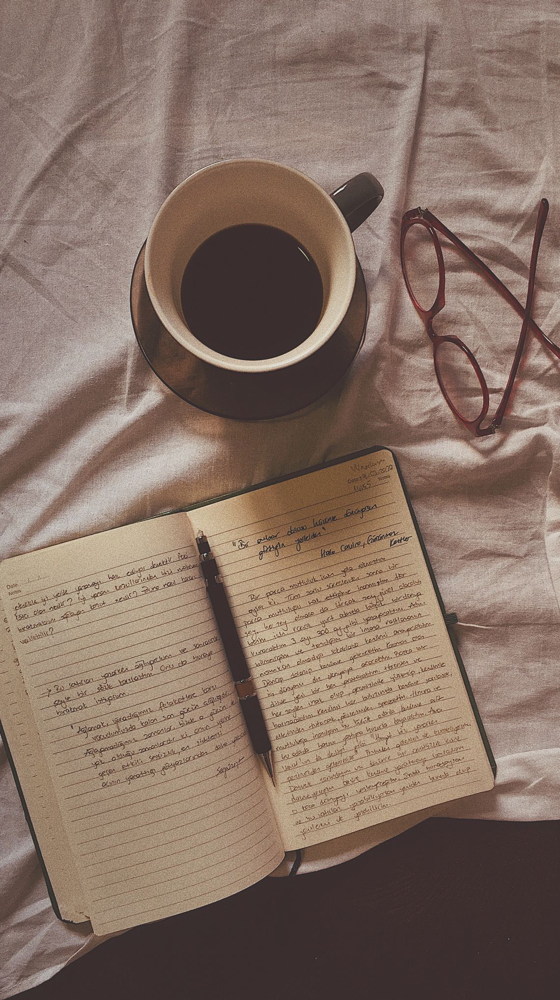 Flat lay of coffee, open notebook, pen, and eyeglasses on a fabric surface.