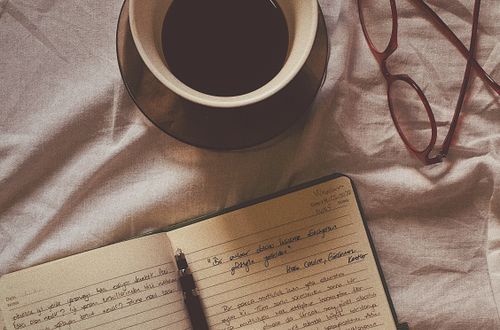 Flat lay of coffee, open notebook, pen, and eyeglasses on a fabric surface.